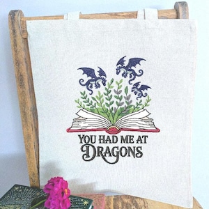 May include: A cream tote bag featuring a dragon design. The design shows three blue dragons above an open book with green leaves. The text "YOU HAD ME AT DRAGONS" is below the book in black script.