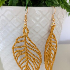 May include: Mustard yellow leaf-shaped earrings with gold-colored hooks. The earrings are made of a textured material, and the leaf design features detailed veining. The earrings are hanging in front of a white pot with green foliage.