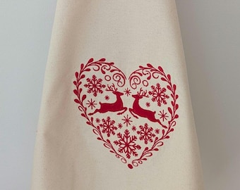 May include: Cream-colored kitchen towel with a red heart-shaped design. The heart contains two reindeer, snowflakes, and decorative flourishes. The towel is made of a soft fabric and is suitable for the holiday season.