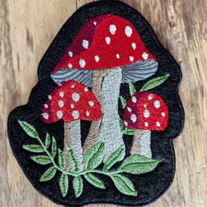 May include: Embroidered patch featuring three red and white mushrooms with white spots on a black background. The mushrooms are surrounded by green leaves.