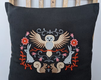 May include: Black throw pillow with embroidered design featuring an owl, rabbits, squirrels, and flowers. The pillow is on a wooden chair.
