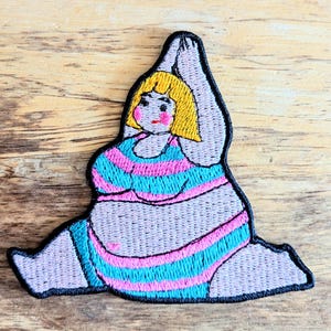May include: Embroidered patch of a person in a striped swimsuit doing a yoga pose with their arm raised above their head. The person has blonde hair and a pink face.