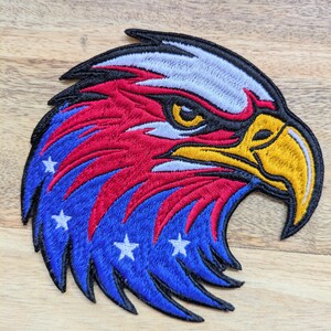 May include: Embroidered patch depicting an eagle's head. The design uses red, white, blue, and yellow thread to create the eagle's features, including feathers and a focused gaze. The blue area features white stars.