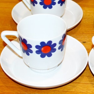 May include: Six white ceramic coffee cups with blue and red floral designs and matching saucers. The cups and saucers have a retro 1970s style.