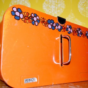 Peut inclure: Boîte en métal orange avec un motif floral et une poignée argentée. La boîte porte une étiquette "Peugeot" sur le fond.