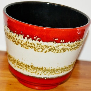 Pode incluir: Um vaso de cerâmica com base e borda vermelhas, uma faixa branca com um padrão texturizado verde-oliva e um interior preto. O vaso tem uma forma ligeiramente angulada e assenta numa superfície de madeira.