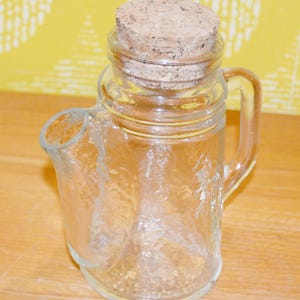 Peut inclure: Une carafe en verre transparent avec une poignée et un bec verseur, surmontée d'un bouchon en liège. La carafe a une surface texturée et est posée sur une surface en bois. Le fond est un mur jaune.
