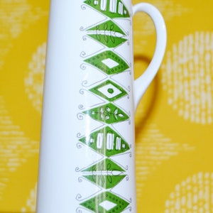 Könnte beinhalten: Eine weiße Keramikkanne mit einem grün-weißen geometrischen Muster. Die Kanne hat einen Henkel und eine hohe, schlanke Form.