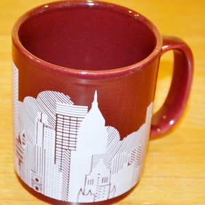 Könnte beinhalten: Eine maronfarbene Keramiktasse mit einer weißen Silhouette einer Skyline. Die Tasse hat einen Henkel auf der rechten Seite.