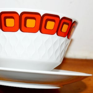 May include: White ceramic bowl and saucer set. The bowl has a repeating pattern of orange and brown squares around the rim. The bowl has a textured, wavy design. The saucer is plain white.