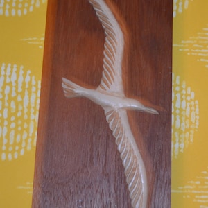 May include: A wooden plaque featuring a carved white bird in flight. The plaque has a brown wood grain and is set against a yellow background.