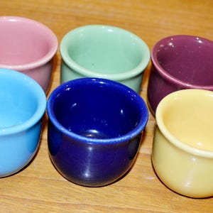 May include: Six small, colorful ceramic bowls in shades of pink, green, purple, blue, navy, and yellow. The bowls are arranged on a wooden surface, showcasing their vibrant colors and rounded shapes. These bowls are ideal for serving dips or small portions.