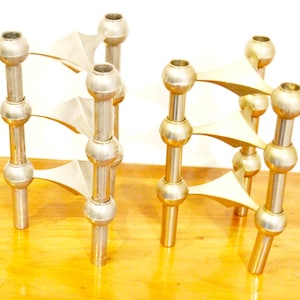 May include: Two sets of vintage metal candle holders. Each holder has a series of vertical posts with spherical accents and triangular supports. The candle holders are made of a shiny, silver-toned metal and sit on a wooden surface.