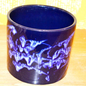 May include: A dark blue ceramic planter with a white and blue abstract design. The planter is cylindrical and has a smooth, glossy finish.