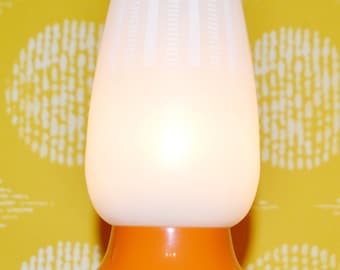 May include: A white and orange table lamp with a textured white shade and a wide orange base. The lamp is turned on and illuminated.