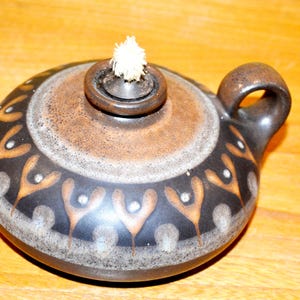 Puede incluir: Una lámpara de aceite de cerámica marrón y negra con una mecha blanca. La lámpara tiene un diseño decorativo de figuras estilizadas alrededor del cuerpo.