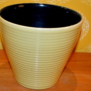 Puede incluir: Una maceta de cerámica amarilla con un interior esmaltado negro. La maceta tiene un diseño acanalado y un fondo redondeado.