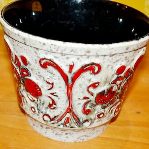 May include: A white ceramic planter with a red and black floral design. The planter has a speckled glaze and a wide, round opening.