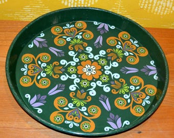 May include: A round, dark green metal tray with a colorful floral design. The pattern features orange, green, white, and purple stylized flowers and swirls on a dark green background. The tray has a slightly raised edge.