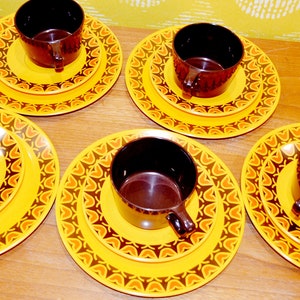 May include: Set of five brown teacups with yellow and orange patterned saucers. The cups and saucers have a geometric design.