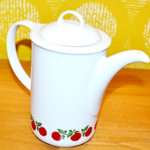 May include: White ceramic teapot with a lid and a curved handle. The teapot features a decorative border of red apples and green leaves. The spout is curved and the lid has a small loop handle. The teapot is on a wooden surface.
