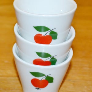 Pode incluir: Três xícaras de ovo de cerâmica branca empilhadas uma em cima da outra. Cada xícara tem um desenho de maçã vermelha com uma folha verde.