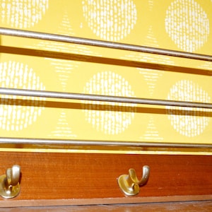 May include: A wooden wall-mounted coat rack with three metal bars and three brass hooks. The rack is mounted on a yellow patterned wall.