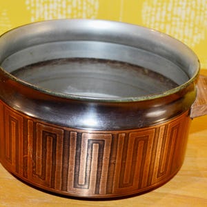 Könnte beinhalten: Ein Vintage-Topf aus kupferfarbenem Metall mit einem geometrischen Design am Boden und Holzgriffen. Der Innenraum ist silberfarben. Der Topf wird wahrscheinlich zum Kochen oder Servieren von Speisen verwendet.