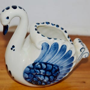 May include: A white ceramic swan-shaped planter with blue accents. The swan features a curved neck, a blue beak, and blue wing details. The planter has a round opening for plants or flowers, and is a decorative item.