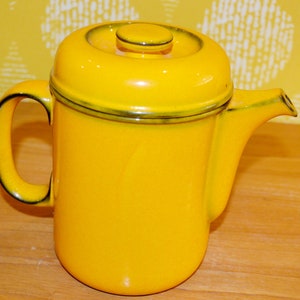 May include: A yellow ceramic coffee pot with a lid and a dark brown trim around the top. The pot has a handle on the left side and a spout on the right side.