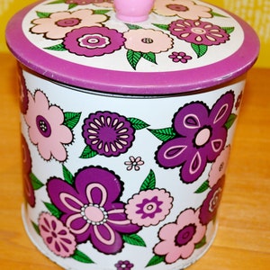 May include: A white metal canister with a purple lid and a pink knob. The canister is decorated with a floral pattern in shades of pink, purple, and green.
