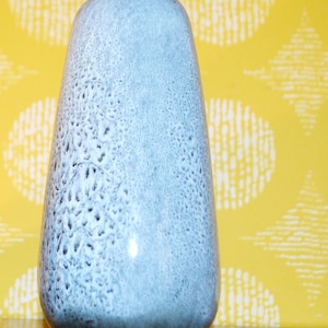 Puede incluir: Un jarrón alto de cerámica azul claro con un esmalte moteado. El jarrón tiene una base redondeada ligeramente acampanada y una abertura estrecha.