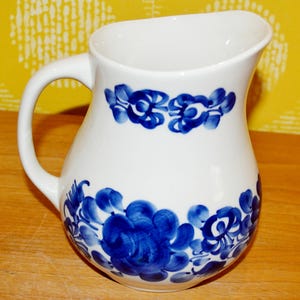 Op de afbeelding: Witte keramische kan met een gebogen handvat en een brede tuit. De kan is versierd met handgeschilderde blauwe bloemmotieven rond de basis en nabij de bovenkant. De kan wordt waarschijnlijk gebruikt voor het serveren van dranken.