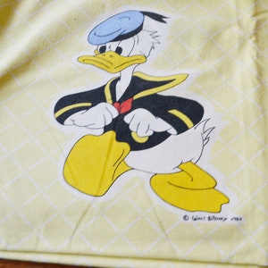 Könnte beinhalten: Ein gelber Stoff mit einem weißen Gittermuster zeigt ein Comicbild von Donald Duck, der einen blauen Matrosenhut und eine schwarz-gelbe Matrosenuniform trägt. Der Text "Walt Disney 1987" ist in der rechten unteren Ecke gedruckt.