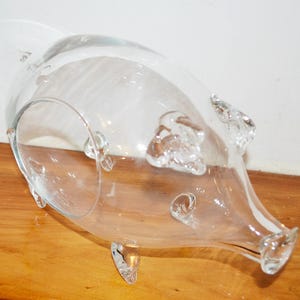 Puede incluir: Un jarrón de vidrio transparente con un diseño único en forma de cerdo. El jarrón tiene una abertura ancha, un cuerpo redondeado y un pico en forma de hocico. Cuenta con pequeños detalles de vidrio transparente que se asemejan a orejas y patas. El jarrón está sobre una superficie de madera.