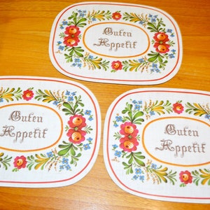 Pode incluir: Três  placemats ovais brancas com uma borda floral vermelha, amarela, verde e azul. O centro de cada placemat tem o texto "Guten Appetit" em escrita cursiva marrom.