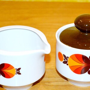 Könnte beinhalten: Ein weißer Keramik-Milchkännchen- und Zuckertopf-Satz mit braunem Deckel. Beide Teile haben ein farbenfrohes geometrisches Design mit orangefarbenen, roten und braunen Akzenten.