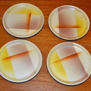 May include: Set of four ceramic coasters with a geometric design. The coasters feature a white background with yellow and brown stripes.