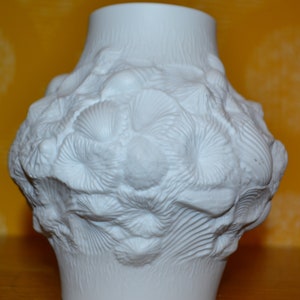 Puede incluir: Jarrón de cerámica blanca con una superficie texturizada que se asemeja a coral o conchas marinas. El jarrón tiene una forma redondeada y una abertura ancha.