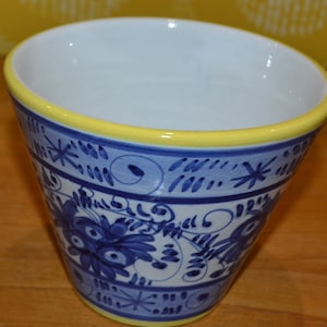 May include: A white ceramic planter with a blue and yellow floral design. The planter has a yellow rim and a blue and white floral pattern.