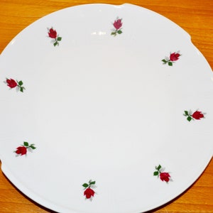 Könnte beinhalten: Ein weißer Keramikteller mit einem gewellten Rand und einem einfachen Design aus roten Rosen mit grünen Blättern.