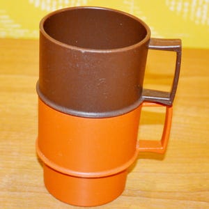 Può includere: Una tazza bicolore marrone e arancione. La sezione superiore è marrone e quella inferiore è arancione. La tazza ha un manico quadrato. La tazza è di plastica ed è probabilmente vintage.