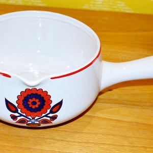 Op de afbeelding: Witte keramische sauspan met een rode rand en een bloemmotief in rood, blauw en wit. De pan heeft een enkele handgreep.