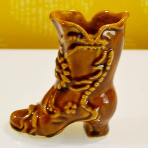 May include: A glossy brown ceramic boot-shaped vase featuring raised relief details including floral patterns and decorative lacing. The object has a scalloped top edge and a distinct heel, with a smooth, reflective finish throughout the entire surface.