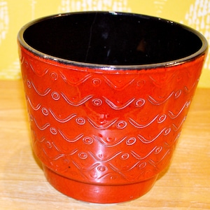 Può includere: Vaso di ceramica rosso con interno nero. L'esterno presenta un motivo a onde ripetute con piccoli cerchi. Il bordo ha un accento argentato. Adatto per piante da interno.