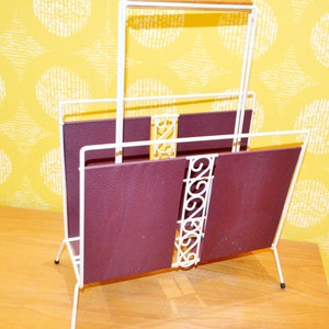 May include: White metal magazine rack with a burgundy leather insert. The rack has a decorative design on the front and a wooden handle on the top.