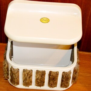 May include: A vintage off-white vanity box with a hinged lid. The box has a rectangular shape with a decorative, furry trim in shades of brown. A small, gold-colored sticker is affixed to the lid. The box is open, revealing a white interior.