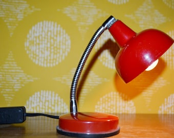 May include: A red desk lamp with a flexible arm and a round base. The lamp has a white light bulb and is plugged into an outlet.