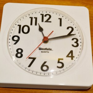 Peut inclure: Une horloge carrée blanche avec des chiffres et des aiguilles noirs. Le cadran de l'horloge est en plastique transparent et l'horloge indique "Westfalia quartz" en bas.