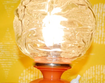 May include: A vintage table lamp with a clear glass globe shade with a textured pattern and an orange base with a gold trim.